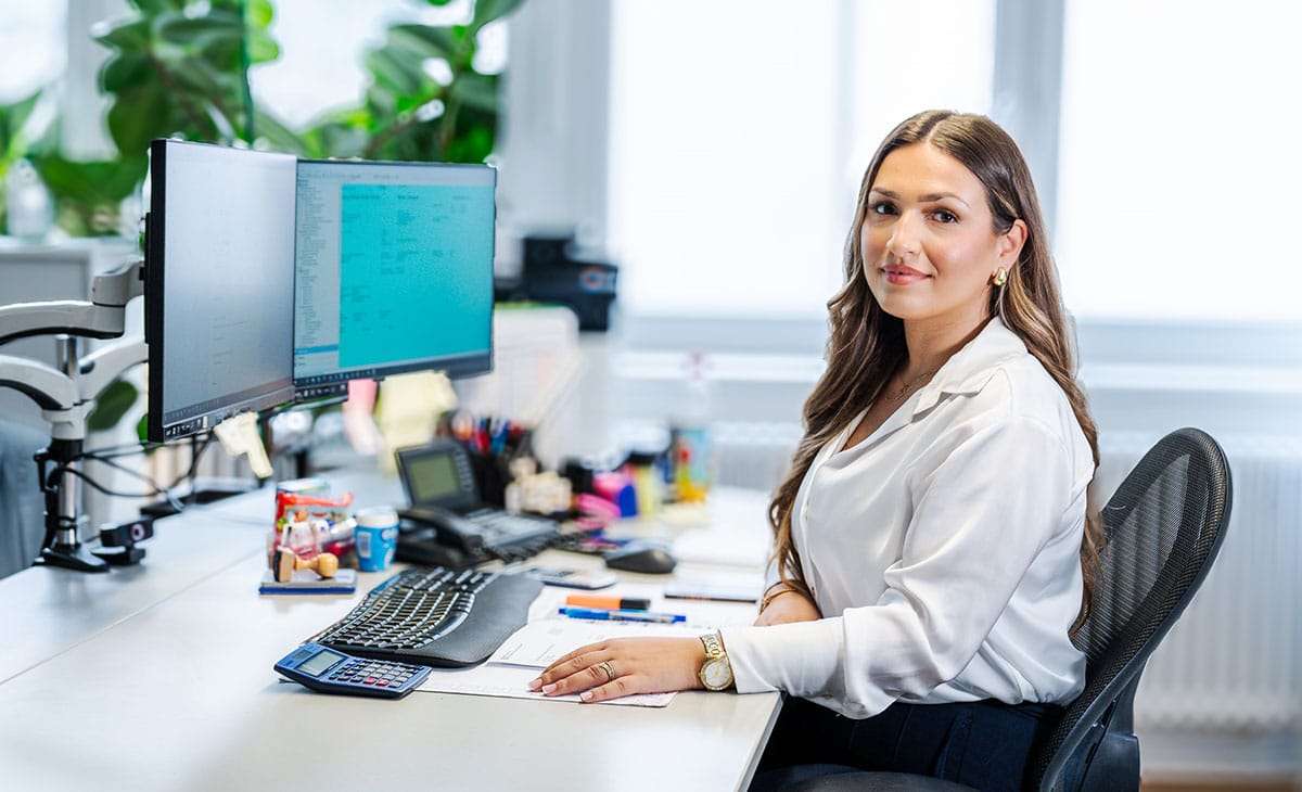 Eine Frau lächelt am Schreibtisch mit zwei Monitoren, umgeben von Büromaterialien.